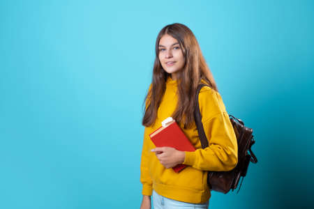 A young student with a backpack looks at a book and reads. Education and training, visiting the library. Young woman in yellow hoodie and jeans on a blue background, copy spaceの写真素材