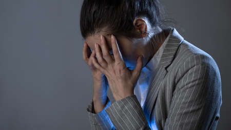 Despair and headache, problems at work and in business, experiencing a crisis. young woman in a business suit standing clutching head, portrait on a gray backgroundの写真素材
