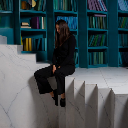 A sad young woman is sitting on a strange staircase against the background of bookshelves. Learning curve, concept. The first failures and loss of motivation.の写真素材