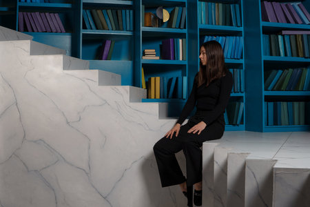 Professional retraining, change of occupation in middle of life. Concept, young woman sitting on surreal staircase changing direction and orientation in space, against the background of bookshelvesの写真素材