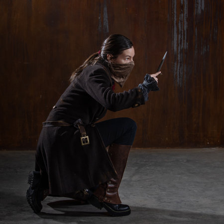 noble robber with a dagger in a squat, preparing to attack. A woman in a retro fantasy outfit on the background of a textured rusty wallの写真素材