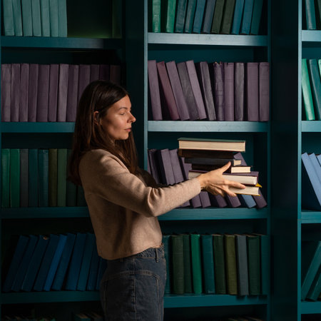 A student in the library, preparing for exams, studying at the university. A young woman holds a stack of heavy textbooks in her handsの写真素材