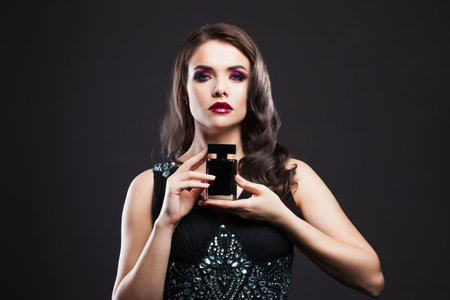 beautiful young woman with bright makeup is holding a bottle of perfume. Gorgeous young brunette in a chic evening look.の写真素材