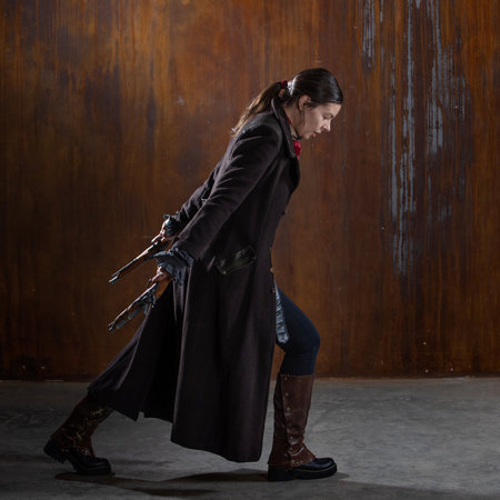 Fantasy steampunk action heroine in a long coat with revolvers in her hands. A woman in a retro fantasy outfit on the background of a textured rusty wallの写真素材