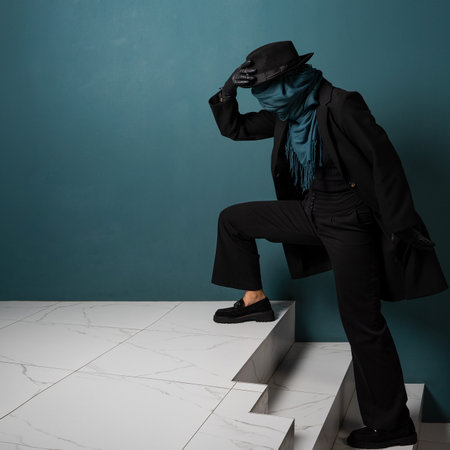 An unknown person, a criminal or a person hiding from the law. Hiding from observation, hiding. A character in a coat and hat with a scarf on his face, sneaks up the stairsの写真素材
