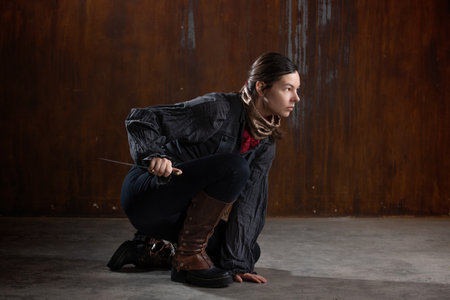 noble robber with a dagger in a squat, preparing to attack. A woman in a retro fantasy outfit on the background of a textured rusty wallの写真素材