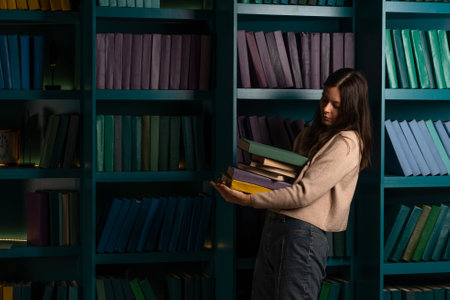 A student in the library, preparing for exams, studying at the university. A young woman holds a stack of heavy textbooks in her handsの写真素材