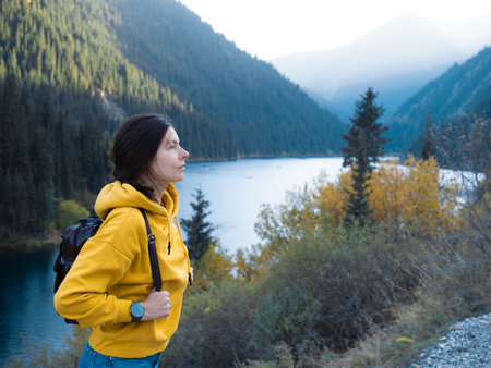 A young female tourist in a yellow hoodie goes hiking with a backpack on her back, picturesque mountain lake in the backgroundの写真素材