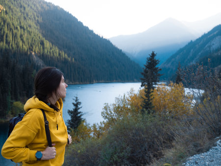 A young female tourist in a yellow hoodie goes hiking with a backpack on her back, picturesque mountain lake in the backgroundの写真素材