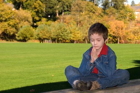 young boy sitting on a wood bench in a park praying or meditating