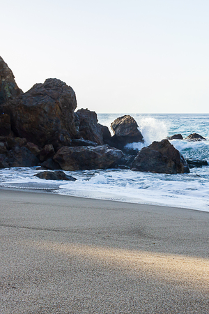 splashing wave breaking on cliff face with rocks near sandy shoreline