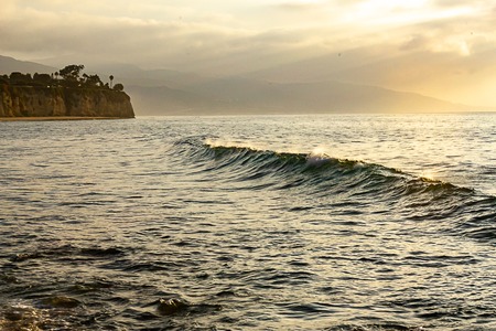 swelling dark wave wall sillouetted against glowing morning ocean expanse with peninsulla and distant hillsの写真素材