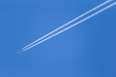 Airplane is flying high in the clear blue sky. Two white condensation trail cross air space.の写真素材