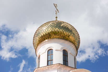 A golden dome with a cross shines in the sun. White tower of Christian church and blue sky with...