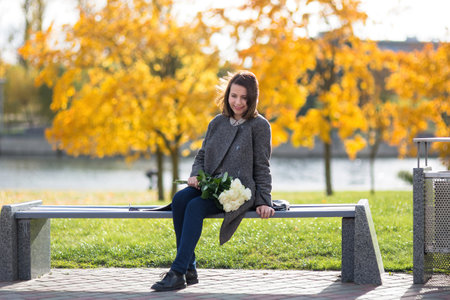 A woman in a coat sits on a bench in a sunny autumn park. She is holding a bouquet of white roses, she smiles and looks down. Hair disheveled by the wind, trees with yellow foliage on the background.の写真素材