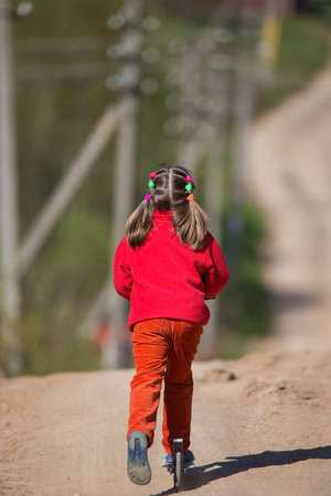 A girl in bright red clothes is riding a scooter down a hill along a dirt country road.の写真素材