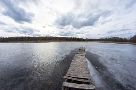 Cold landscape of a frozen lake with a long wooden pier. Cloudy sky and forest on the far side of the pond.の写真素材