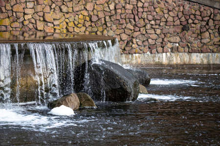 Small waterfall in the city park. Jets of water fall on stones, spray is flying. There is a natural stone wall above the waterfall.の写真素材