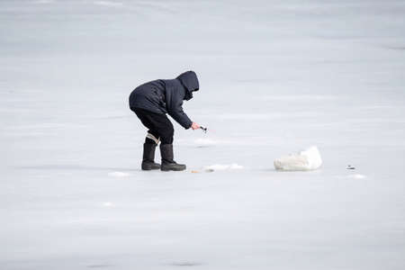 A lone fisherman is bent over a hole in the ice and is trying to catch a fish.の写真素材