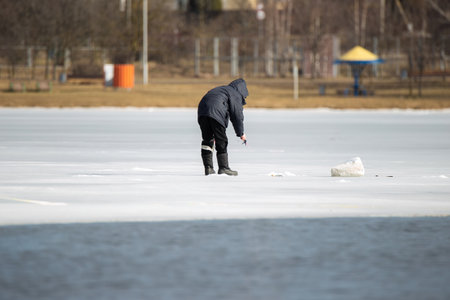 A lone fisherman is bent over a hole in the ice and is trying to catch a fish dangerously close to the edge of the thin melting ice.の写真素材