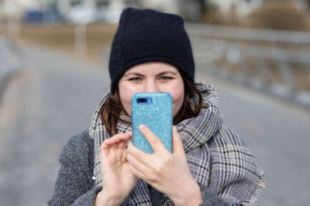 A woman in warm clothes stands in the street and holds a smartphone in a bright blue case in front of her face. A woman takes a selfie in the street.の写真素材