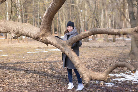 A woman in warm clothes and white sneakers stands behind a fallen tree in a sunny city park in early spring.の写真素材