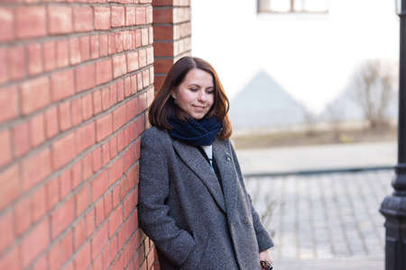 A beautiful woman with colorful brown hair in a gray coat and a blue scarf stands against a red brick wall. She is leaning against the wall, smiling and looking down.の写真素材