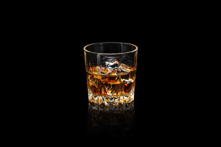 Glass of whiskey with ice cubes on a dark reflective surface isolated on black.