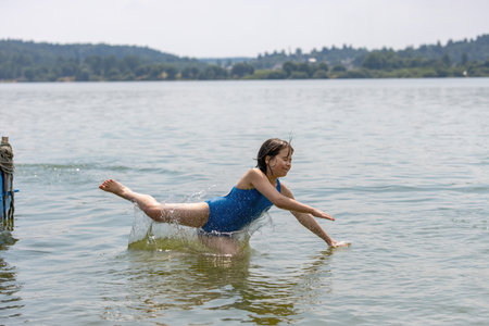 Teenage girl in blue swimsuit bathes in calm cool lake water in hot summer, raising a lot of splashesの写真素材
