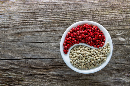 A halved porcelain bowl filled with white and red peppercorns on an aged rough board.の写真素材
