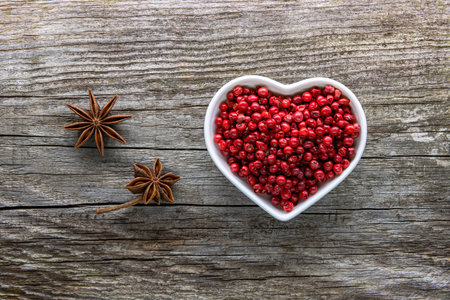 A heart-shaped porcelain bowl filled with red peppercorns on an aged rough board. Near the...