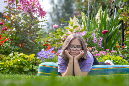 A sleepy girl in glasses lies with her eyes closed, her head resting on a mattress on the lawn in a summer blooming gardenの写真素材