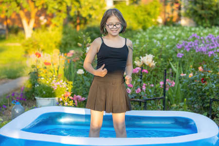 A girl with beautiful multi-colored hair in glasses stands in cool water in an inflatable pool in an evening hot summer gardenの写真素材