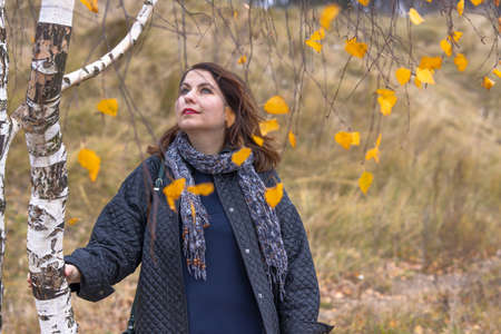 A woman with long hair and red lipstick on her lips stands among the branches and yellow leaves of a birchの写真素材