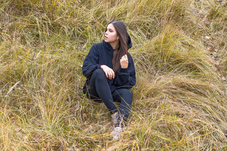 A beautiful girl with long hair in black clothes sits on a hillside on the autumn grass and looks to the sideの写真素材