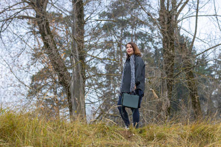 A woman with a handbag in autumn clothes walks on the grass in a deserted place against the background of autumn treesの写真素材