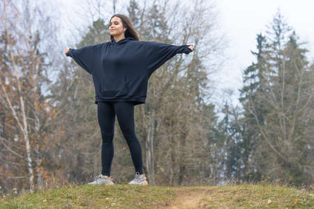 A beautiful girl with long hair with wireless headphones in her ears in black clothes stands with her arms outstretched on a hill on the grass and smilesの写真素材