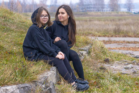 Two beautiful girls with long hair in black clothes sit together on the grass on the outskirts of the city.の写真素材