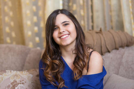 Young smiling beautiful girl with long hair sits on a soft sofa in a cozy homeの写真素材