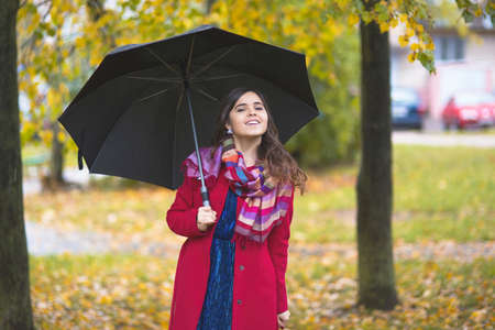 Young beautiful girl in a red coat stands with a large umbrella in an autumn park among yellow leavesの写真素材