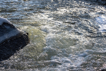 The rapid flow of the river breaks on a large stone covered with ice. The water frosts and sparkles in the spring sun.の写真素材