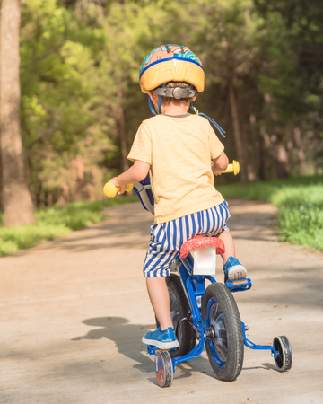 Rear view of Asian toddler boy riding tricycle at nature park near Dallas, Texas, USA. Healthy kid cycling with helmet, short and sneaker on park concrete pathwayの写真素材