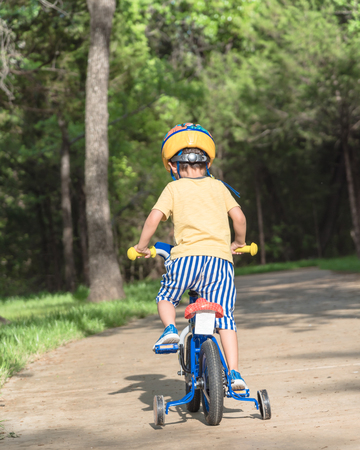 Rear view of Asian toddler boy riding tricycle at nature park near Dallas, Texas, USA. Healthy kid cycling with helmet, short and sneaker on park concrete pathwayの写真素材
