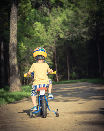 Vintage tone rear view of Asian toddler boy riding tricycle at nature park near Dallas, Texas, USA. Healthy kid cycling with helmet, short and sneaker on park concrete pathwayの写真素材