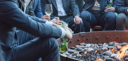 Group of Caucasian businessmen in formal dress gathering over the round fire pit after work in Chicago. Business people in formal dress with watches, leather shoes drinking beer bottle, wine glassesの写真素材