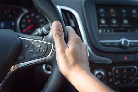 Close-up Asian male hand holding modern car steering wheel with audio control buttons. Voice and...