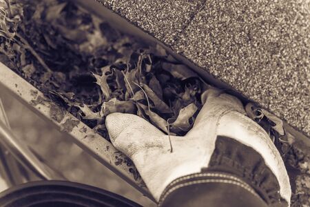 Vintage tone close-up man hand with gloves on ladder cleaning house gutter from leaves and dirt. Roof gutter cleaning near shingles roof in summer timeの写真素材