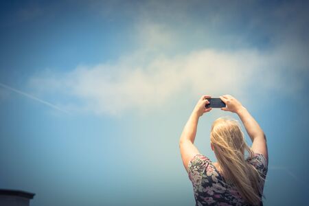 Lookup view of young Caucasian lady taking photo using her smart phone. Low angle blonde girl with...
