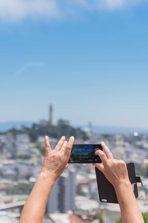 Rear view of tourist using smart phone taking photo of Coit Tower in San Francisco, California. The other hand of traveler holding the leather cell phone flip case.の写真素材