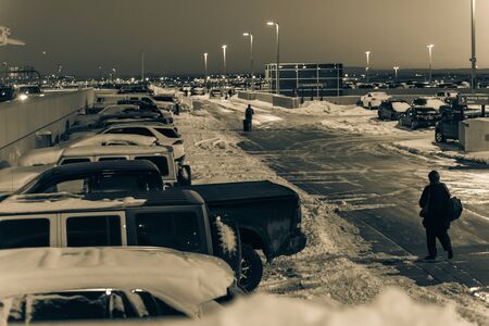 Rear view travelers in warm clothes walking with luggage at terminal parking of Denver International Airport (DIA) in frosty cold autumn sunset. Busy lot with snowdrifts, severe weather conditionの写真素材